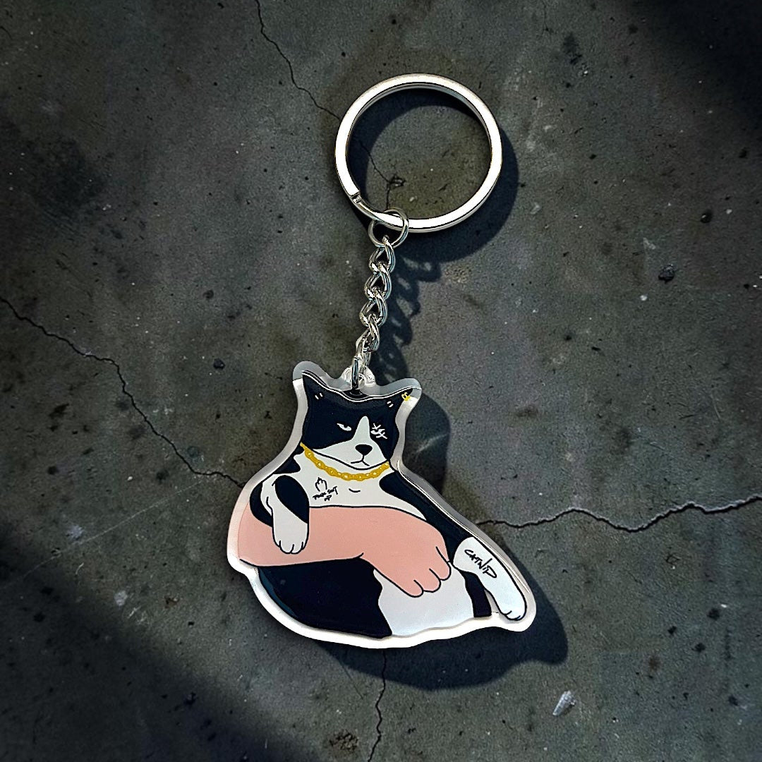 A metal keychain featuring a cartoon cat character with black, white, pink, and yellow details, resting on a dark textured concrete surface.